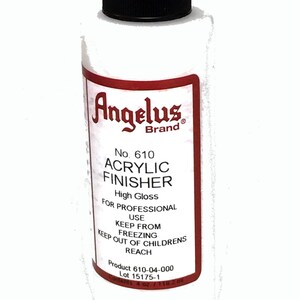 May include: A white bottle of Angelus Brand No. 610 Acrylic Finisher, a high gloss sealant for professional use. The bottle has a black cap and a red label with white text. The label reads "Keep from freezing. Keep out of children's reach. Product 610-04-000. Lot 15175-1. Contents 4 oz / 118.2 ml."