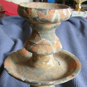 May include: An antique, weathered candlestick holder with a tiered design. The ceramic piece features a wide base, a central dish, and a top cup, all adorned with faded orange and gray paint. The overall height is approximately 7 inches.