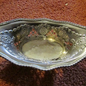 Puede incluir: Plato ovalado plateado con detalles florales y de volutas ornamentados. El plato tiene un diseño calado en el centro y un borde decorativo en forma de cuerda. El objeto es probablemente de metal y está sobre una superficie marrón.