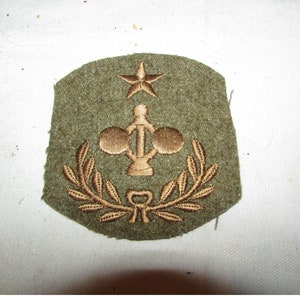 May include: Olive green military patch with embroidered gold-colored star, emblem, and laurel wreath design. The patch has a slightly curved top and a cut edge. The emblem features a central vertical element with two circular shapes on either side.