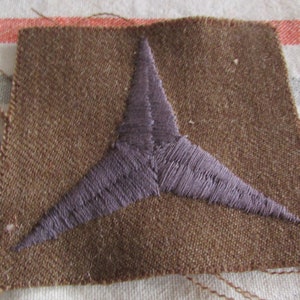 May include: A brown fabric patch with a purple embroidered three-pointed star design. The patch has a square shape with visible thread edges. The background is a striped fabric.
