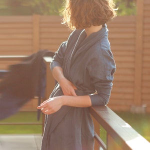 May include: A woman wearing a blue linen robe stands by a wooden railing. The robe has a relaxed fit and a shawl collar. The woman's hair is curly and brown.