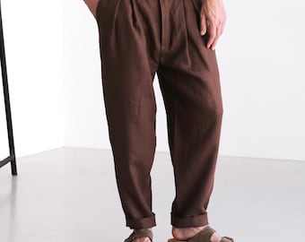 May include: Brown pleated trousers with a button closure, paired with brown sandals. The pants have a relaxed fit. A black side table with a black vase and a magazine is in the background. The image has an updated sizing chart.