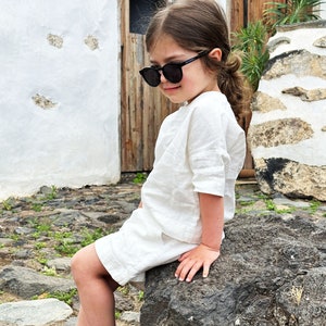 May include: A young person wearing a white linen shirt and black sunglasses sits on a large rock. The person is barefoot and has their hair pulled back in a ponytail.