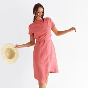 May include: A woman wearing a coral pink linen wrap dress with short sleeves and a belt. She is holding a straw hat in her right hand.