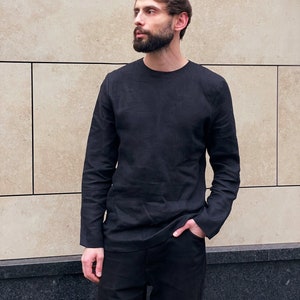 May include: A man wearing a black long-sleeved shirt with a crew neck and black trousers. He is standing in front of a tiled wall.