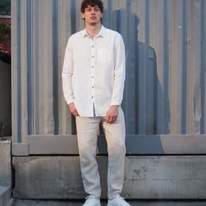 Pu&ograve; includere: Un uomo indossa una camicia di lino bianca con colletto abbottonato e una tasca sul petto, pantaloni di lino beige e scarpe da ginnastica bianche. &Egrave; in piedi davanti a un muro di metallo grigio.