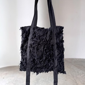 Peut inclure: Sac fourre-tout noir avec de longues anses. Le corps du sac pr&eacute;sente un motif textur&eacute; en tissu d&eacute;chiquet&eacute;. Les anses sont longues et fabriqu&eacute;es dans le m&ecirc;me mat&eacute;riau noir. Le sac est tenu par une main, mais l'accent est mis sur le sac lui-m&ecirc;me.