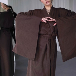 May include: A brown linen robe with long sleeves and a tie belt. The robe is open in the front, revealing a glimpse of the person's chest. The person is wearing a necklace with a pendant.