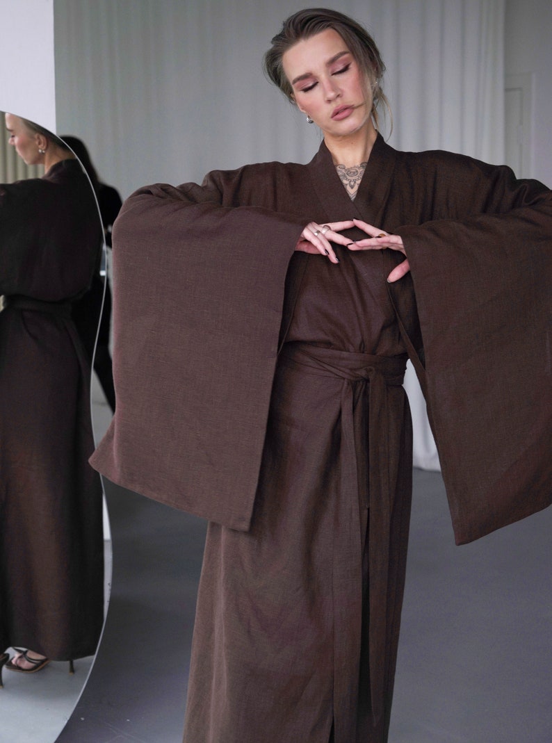 May include: A brown linen robe with long sleeves and a tie belt. The robe is open in the front, revealing a glimpse of the person's chest. The person is wearing a necklace with a pendant.