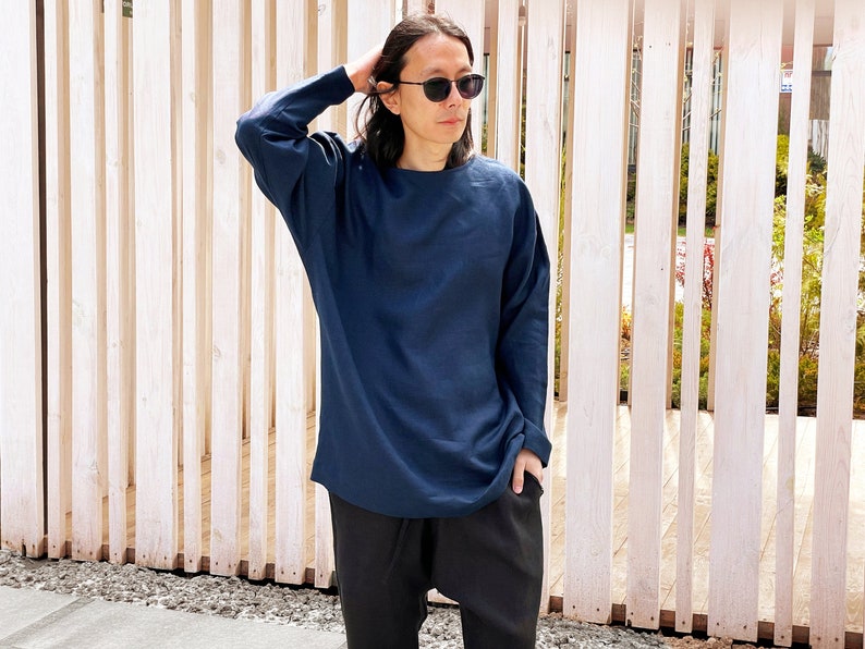 May include: A person wearing a dark blue long-sleeved shirt and black pants. The person is standing in front of a wooden slat wall.