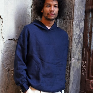 Pode incluir: Um moletom azul marinho com capuz e mangas compridas. O modelo está vestindo uma camiseta rosa claro e calças creme. O fundo é uma parede de pedra desgastada. Um círculo branco com o texto "UPDATED SIZING CHART*" está no canto inferior direito.
