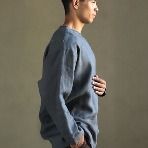 May include: A man wearing a gray linen shirt with long sleeves and a relaxed fit. He is wearing dark brown pants and has his hand in his pocket.