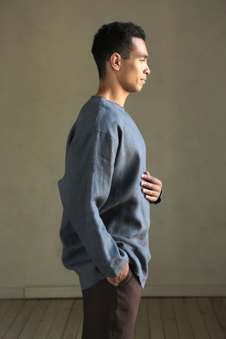 May include: A man wearing a gray linen shirt with long sleeves and a relaxed fit. He is wearing dark brown pants and has his hand in his pocket.