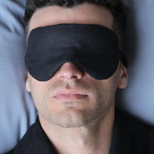 K&ouml;nnte beinhalten: Eine Person tr&auml;gt eine schwarze Schlafmaske, w&auml;hrend sie im Bett liegt. Die Schlafmaske ist aus einem weichen, bequemen Stoff gefertigt und soll Licht abhalten.