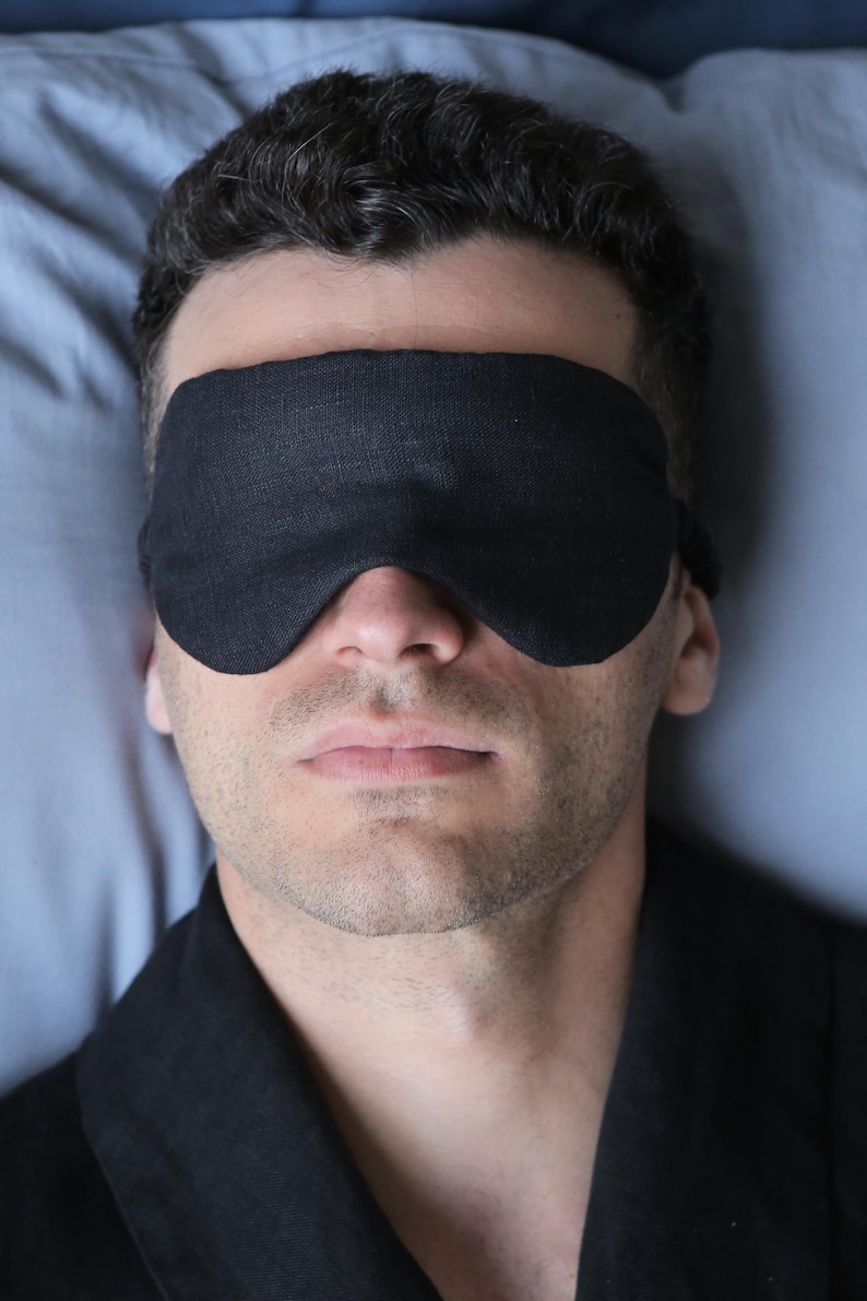K&ouml;nnte beinhalten: Eine Person tr&auml;gt eine schwarze Schlafmaske, w&auml;hrend sie im Bett liegt. Die Schlafmaske ist aus einem weichen, bequemen Stoff gefertigt und soll Licht abhalten.