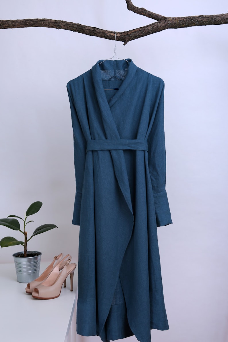 May include: A teal blue linen wrap dress with a long sleeve and a tie belt. The dress is hanging on a wooden branch.