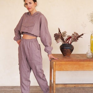 May include: A mauve linen hoodie and matching pants with a drawstring waist and cargo pockets. The model is standing next to a wooden table with a vase of dried flowers.