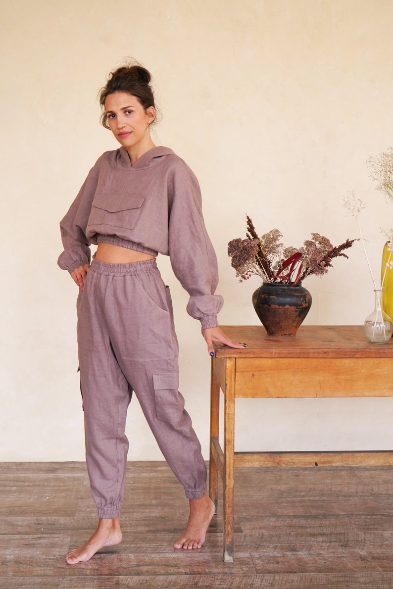 May include: A mauve linen hoodie and matching pants with a drawstring waist and cargo pockets. The model is standing next to a wooden table with a vase of dried flowers.