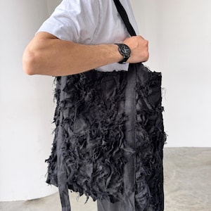 Peut inclure: Sac fourre-tout noir avec un design en tissu textur&eacute; et d&eacute;chiquet&eacute;. Le sac a de longues sangles et une forme rectangulaire. Le sac est port&eacute; sur l'&eacute;paule. Le sac est fait d'un mat&eacute;riau sombre.