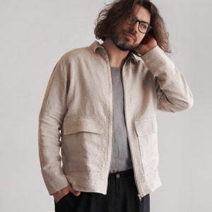 Puede incluir: Un hombre con una chaqueta de lino beige claro con cierre de cremallera y dos bolsillos grandes. Tambi&eacute;n lleva pantalones negros.