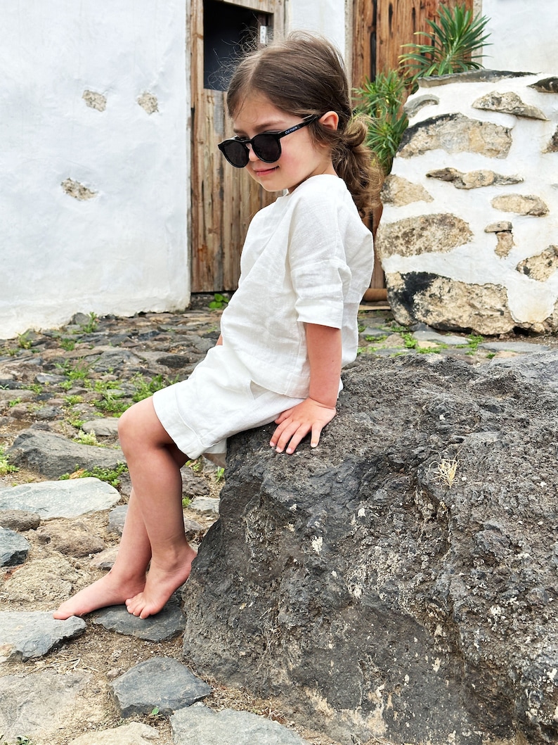 May include: A young person wearing a white linen shirt and black sunglasses sits on a large rock. The person is barefoot and has their hair pulled back in a ponytail.