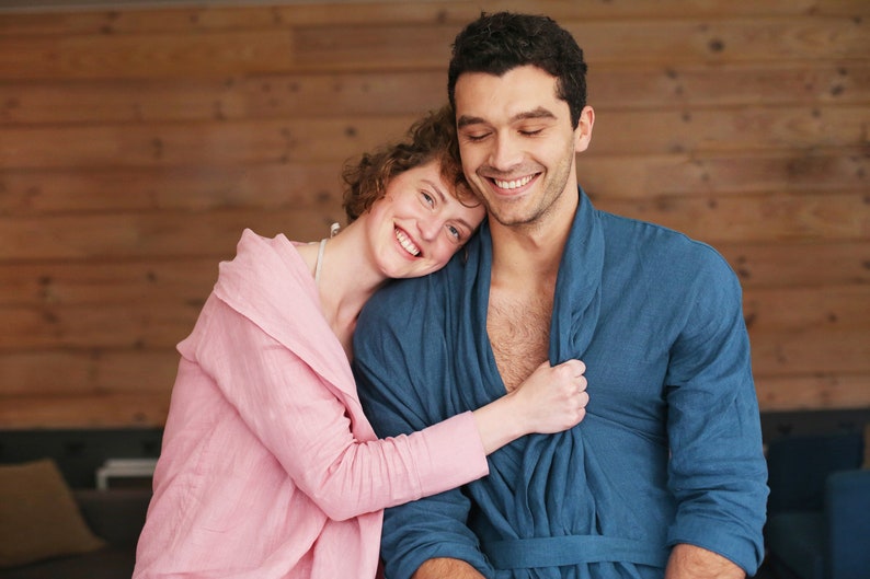 Pode incluir: Um casal em um abra&ccedil;o amoroso, vestindo roup&otilde;es combinando azul e rosa. A mulher est&aacute; com a cabe&ccedil;a apoiada no ombro do homem, e ambos est&atilde;o sorrindo.