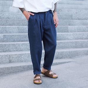 May include: Navy blue pleated pants with a relaxed fit. The pants are cuffed at the ankles and paired with brown sandals. The person is wearing a white shirt. The image is taken outdoors on a set of stairs.