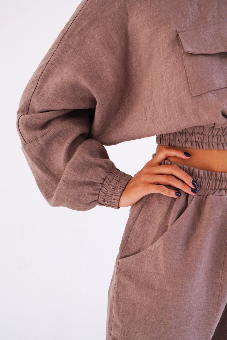 May include: A brown linen two-piece set with a cropped top and pants. The top has a relaxed fit with long sleeves and a drawstring waist. The pants have a relaxed fit with pockets and a drawstring waist.