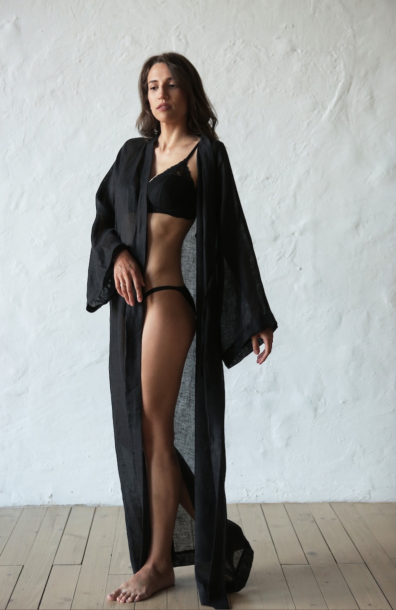 May include: A woman wearing a black linen robe with a black lace bra and black underwear. The robe is open in the front, revealing her legs and torso. She is standing on a wooden floor in front of a white wall.