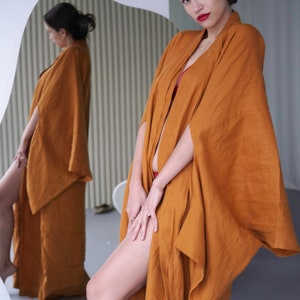 May include: A woman wearing a long, rust-colored linen robe with wide sleeves. The robe is open in the front, revealing a red undergarment. The woman is standing in front of a mirror, looking at the camera.