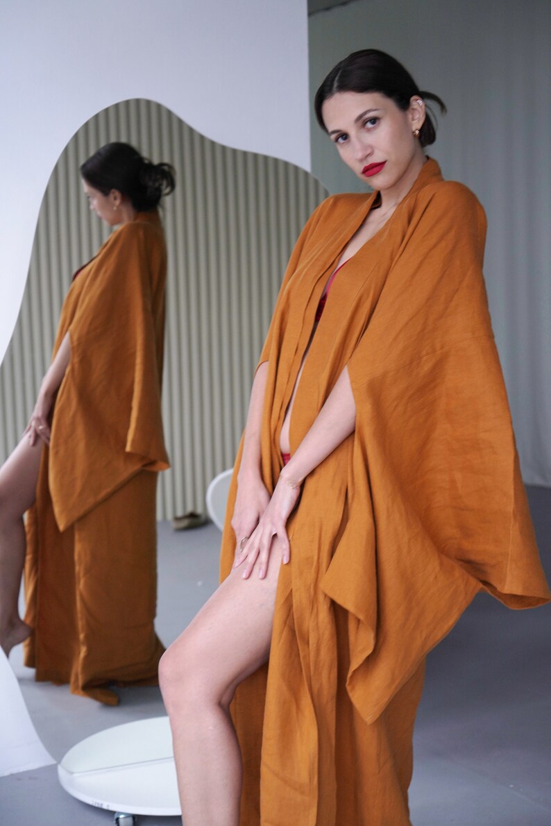 May include: A woman wearing a long, rust-colored linen robe with wide sleeves. The robe is open in the front, revealing a red undergarment. The woman is standing in front of a mirror, looking at the camera.