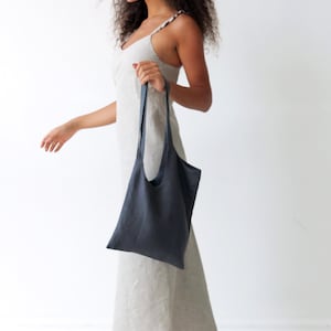 May include: A woman wearing a light-coloured linen dress is walking while holding a dark grey linen tote bag.