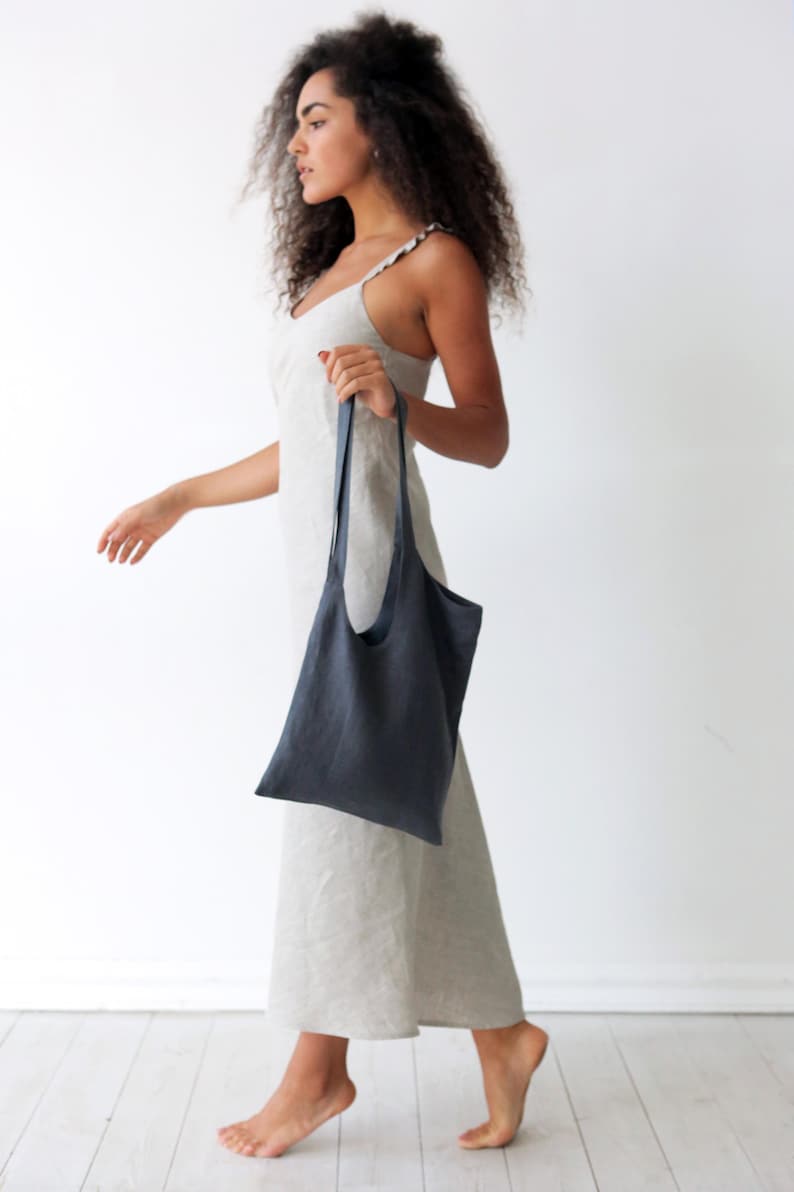 May include: A woman wearing a light-coloured linen dress is walking while holding a dark grey linen tote bag.