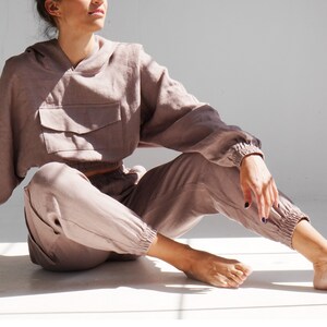 May include: A mauve-colored linen hoodie and matching pants. The hoodie has a large pocket on the front and a drawstring hood. The pants have an elastic waistband and cuffed legs.
