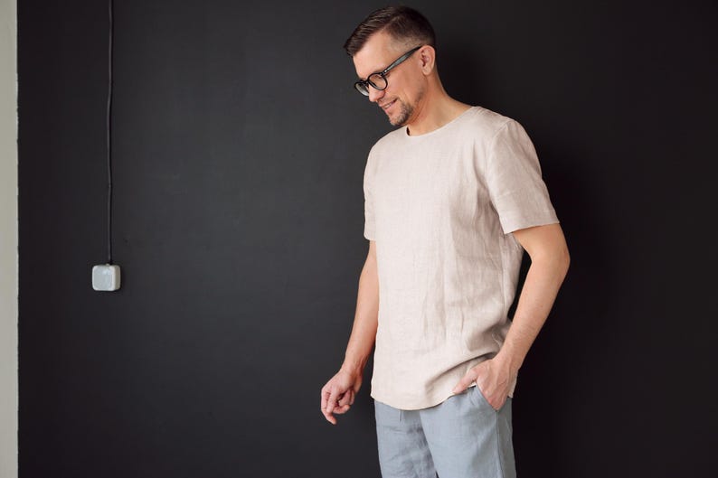 May include: A man wearing a light beige linen shirt with a crew neck and short sleeves. He is standing in front of a black wall.