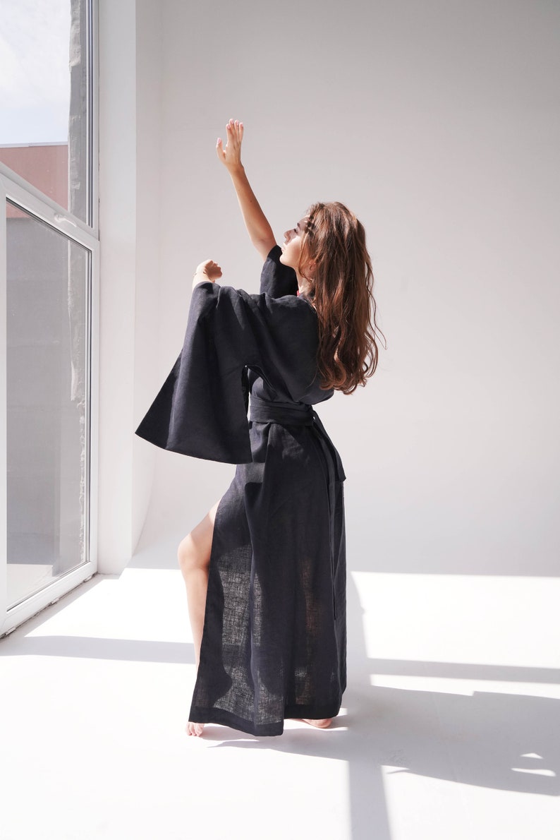 May include: A woman wearing a black linen robe with long sleeves and a sash. The robe has a side slit and the woman is standing with her arm raised.
