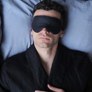 K&ouml;nnte beinhalten: Eine Person mit einer schwarzen Schlafmaske und einem schwarzen Morgenmantel liegt im Bett. Die Person hat die Augen geschlossen und die H&auml;nde auf der Brust liegen.