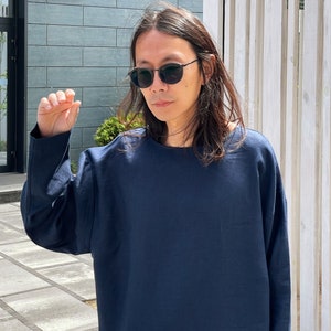 May include: A person wearing a dark blue long-sleeved shirt and black sunglasses. The person is standing in front of a building with a sign that says "2D".