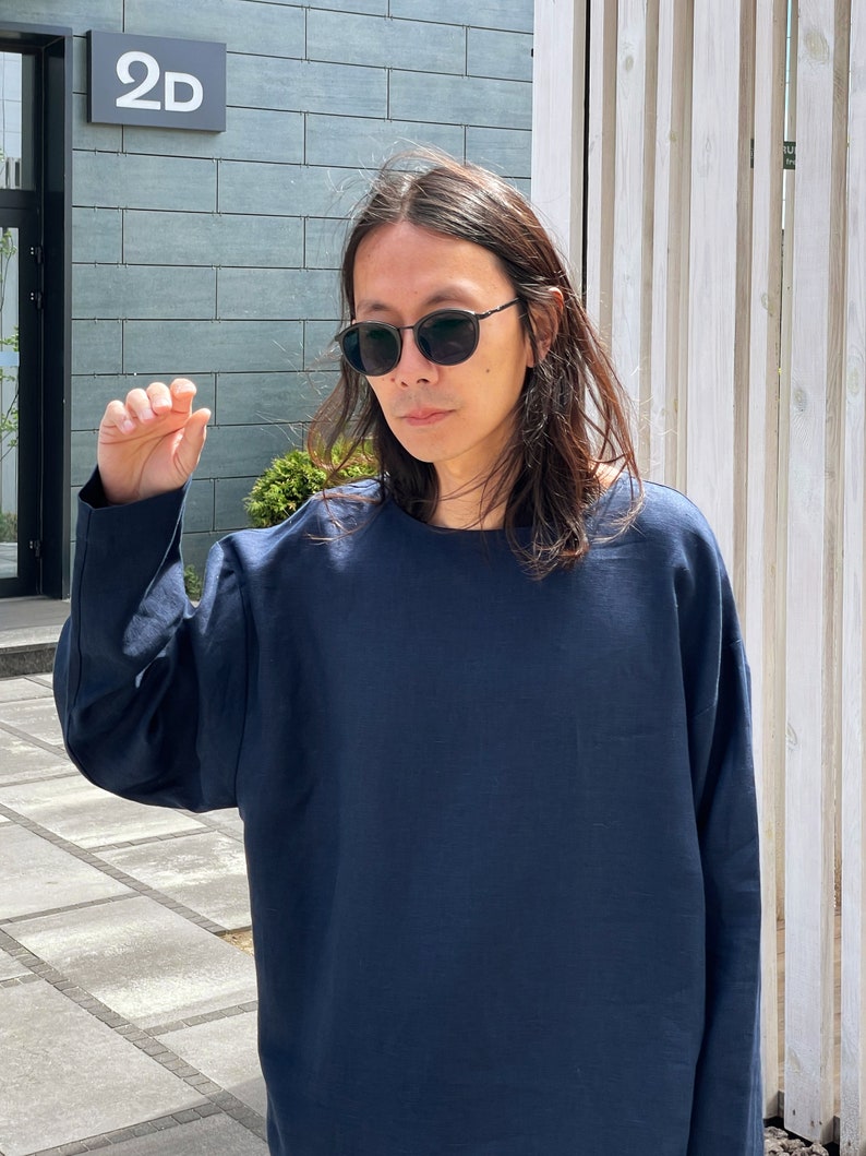 May include: A person wearing a dark blue long-sleeved shirt and black sunglasses. The person is standing in front of a building with a sign that says "2D".
