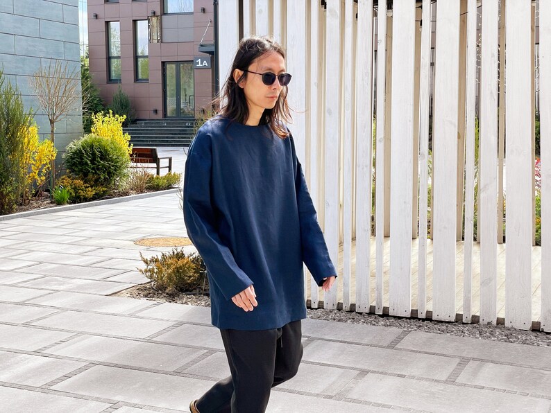 May include: A person wearing a dark blue long-sleeved shirt and black pants. The person is wearing sunglasses and walking outside.
