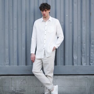 Pu&ograve; includere: Un uomo indossa una camicia di lino bianca e pantaloni di lino beige. &Egrave; in piedi davanti a un muro di metallo ondulato grigio e indossa scarpe da ginnastica bianche.