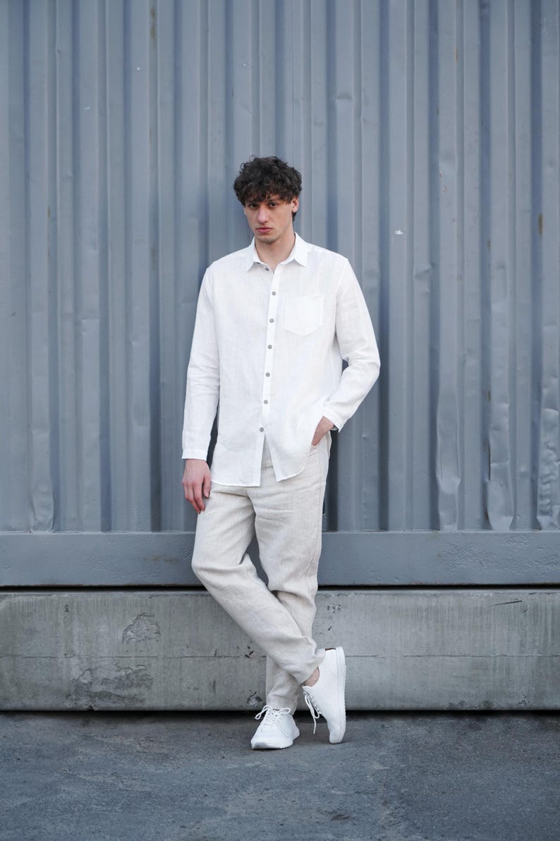 Pu&ograve; includere: Un uomo indossa una camicia di lino bianca e pantaloni di lino beige. &Egrave; in piedi davanti a un muro di metallo ondulato grigio e indossa scarpe da ginnastica bianche.