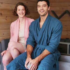 Pode incluir: Um casal em uma sala de estar, vestindo roup&otilde;es combinando. O homem est&aacute; vestindo um roup&atilde;o azul e a mulher um roup&atilde;o rosa. Eles est&atilde;o sentados em um sof&aacute; e sorrindo para a c&acirc;mera.