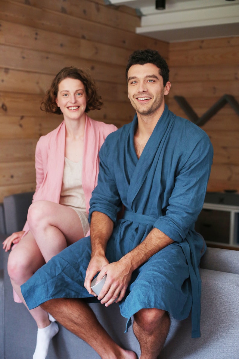 Pode incluir: Um casal em uma sala de estar, vestindo roup&otilde;es combinando. O homem est&aacute; vestindo um roup&atilde;o azul e a mulher um roup&atilde;o rosa. Eles est&atilde;o sentados em um sof&aacute; e sorrindo para a c&acirc;mera.