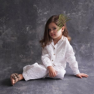 Pu&ograve; includere: Una bambina con una camicia e pantaloni di lino bianco &egrave; seduta su uno sfondo grigio testurizzato. Ha una felce nei capelli e guarda la telecamera.