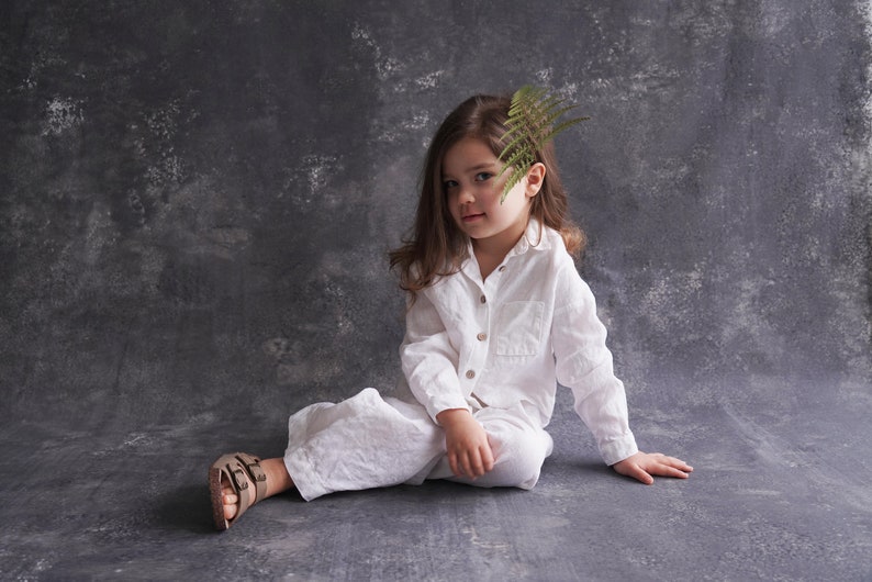 Pu&ograve; includere: Una bambina con una camicia e pantaloni di lino bianco &egrave; seduta su uno sfondo grigio testurizzato. Ha una felce nei capelli e guarda la telecamera.