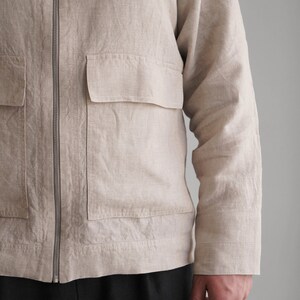 Puede incluir: Una chaqueta de lino beige con cierre de cremallera y un gran bolsillo de parche en la parte delantera. La chaqueta tiene mangas largas y un ajuste holgado.
