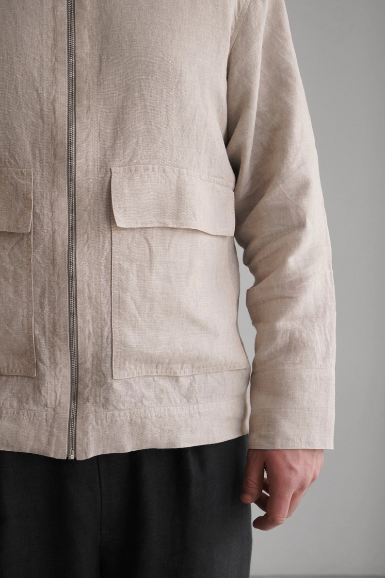 Puede incluir: Una chaqueta de lino beige con cierre de cremallera y un gran bolsillo de parche en la parte delantera. La chaqueta tiene mangas largas y un ajuste holgado.