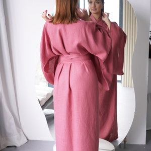 May include: A long, pink linen robe with a wide sash and long sleeves. The robe is tied at the waist and has a relaxed fit.